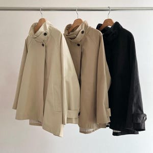 May include: Three jackets hanging on a metal rod. The jackets are in beige and black. The beige jackets have a high collar with buttons and a pocket. The black jacket has a high collar with buttons.
