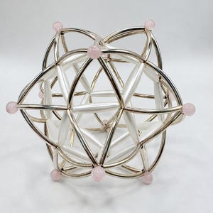 Puede incluir: Una escultura geométrica construida con varillas cilíndricas de color plata metálico y blanco dispuestas en una compleja red en forma de estrella. Los vértices de la estructura están acentuados con pequeñas cuentas esféricas lisas de color rosa pálido.
