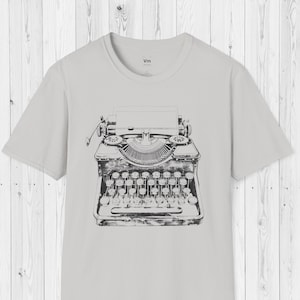 以下が含まれることがあります： 前面にヴィンテージタイプライターの白黒イラストが描かれたグレーのTシャツです。