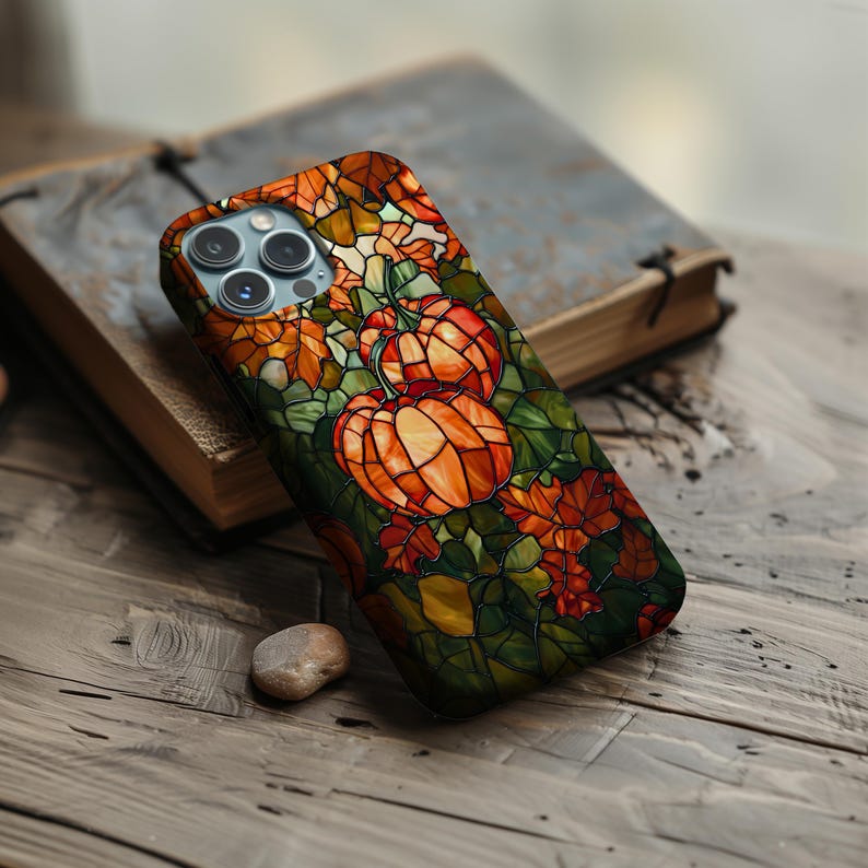 Puede incluir: Una funda para tel&eacute;fono con un dise&ntilde;o de vitral que presenta calabazas y hojas de oto&ntilde;o en tonos naranja, verde y marr&oacute;n.