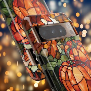Puede incluir: Una funda para tel&eacute;fono con un dise&ntilde;o de vitral que presenta calabazas y hojas en tonos naranja, verde y marr&oacute;n. La funda tiene una protuberancia de c&aacute;mara elevada con una lente de c&aacute;mara negra.