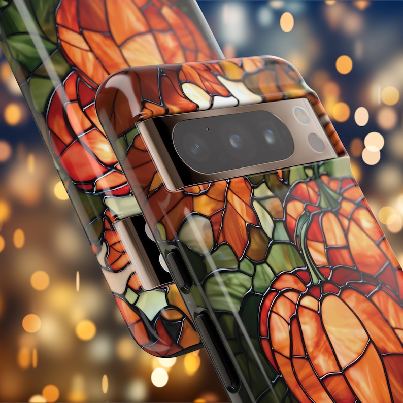 Puede incluir: Una funda para tel&eacute;fono con un dise&ntilde;o de vitral que presenta calabazas y hojas en tonos naranja, verde y marr&oacute;n. La funda tiene una protuberancia de c&aacute;mara elevada con una lente de c&aacute;mara negra.