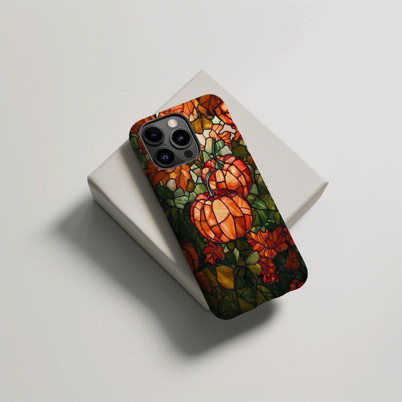 Puede incluir: Una funda para tel&eacute;fono con un dise&ntilde;o de vitral con calabazas naranjas y hojas verdes. La funda es negra y tiene un recorte para la c&aacute;mara del tel&eacute;fono.