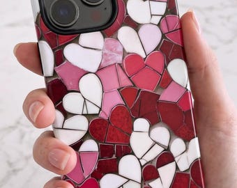 Puede incluir: Una funda para smartphone con un patrón de mosaico de corazones en tonos rojo, rosa y blanco. El diseño consiste en pequeñas piezas irregulares dispuestas para formar motivos de corazones en toda la superficie trasera de la funda.