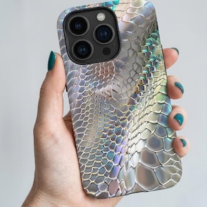 May include: A smartphone case featuring a repeating, iridescent scale pattern in shades of silver, white, and teal. The design mimics a reptile skin texture with a metallic, reflective finish that shifts in color across the surface of the case.