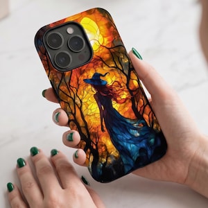 May include: A smartphone case featuring a stained glass style illustration of a figure in a blue dress and hat standing in a forest. The background displays a vibrant orange and yellow sunset sky behind dark tree silhouettes, with a triple camera lens.