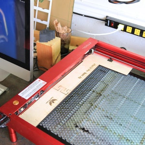 Puede incluir: Cortadora l&aacute;ser roja con una cama de corte de panal y una tabla de madera en su interior. La cortadora l&aacute;ser est&aacute; sobre una mesa junto a un ordenador y un teclado.