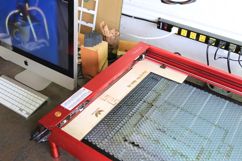 Puede incluir: Cortadora l&aacute;ser roja con una cama de corte de panal y una tabla de madera en su interior. La cortadora l&aacute;ser est&aacute; sobre una mesa junto a un ordenador y un teclado.