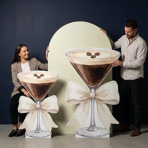 Op de afbeelding: Twee grote, decoratieve martini-glazen uitsparingen. Elk heeft een koffiekleurige drank met koffiebonen erop en een crèmekleurige strik. De uitsparingen worden ondersteund door een voet en bevinden zich voor een donkerblauwe achtergrond.