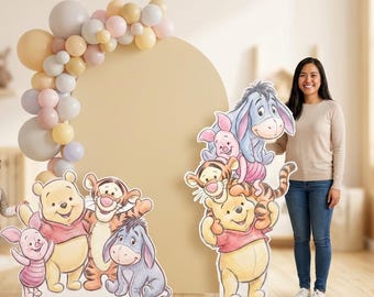 May include: A party scene featuring Winnie the Pooh and friends. The scene includes cardboard cutouts of the characters, a balloon arch, and a large neutral-colored backdrop. A child and an adult are standing near the cutouts.