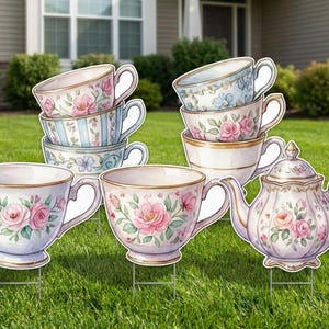 Puede incluir: Señales decorativas de jardín con tazas de té y una tetera. Las tazas y la tetera son blancas con diseños florales en rosa, azul y verde. Las señales están colocadas sobre estacas metálicas en un jardín.
