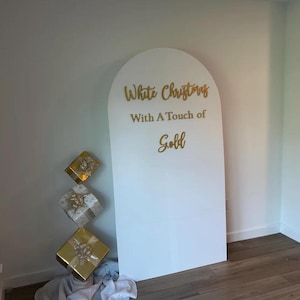 May include: White arch backdrop with the text "White Christmas with a Touch of Gold" in gold lettering. Three gold gift boxes are stacked in front of the backdrop.