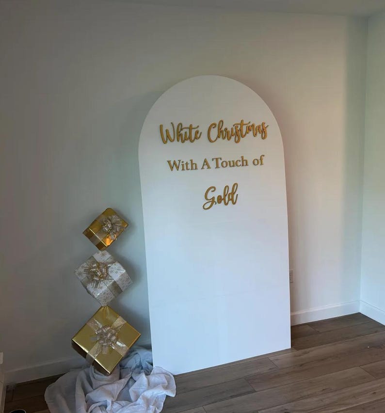 May include: White arch backdrop with the text "White Christmas with a Touch of Gold" in gold lettering. Three gold gift boxes are stacked in front of the backdrop.