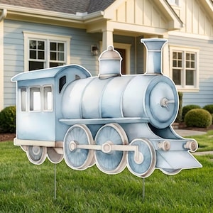 May include: A light blue, watercolour-style train garden ornament. The train features a classic design with a smokestack, wheels, and a passenger carriage. It is positioned on a green lawn in front of a house.