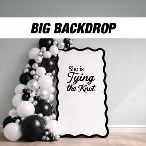 May include: A black and white backdrop with the text "BIG BACKDROP" at the top. A balloon arch in black, white, and silver surrounds a white sign with the text "She is Tying the Knot".