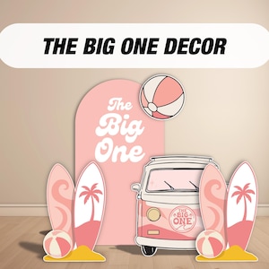 May include: Pink and white beach-themed party decorations. The image features a pink van, surfboards, beach balls, and a backdrop with the text "The Big One". The decorations are set on a wooden floor.