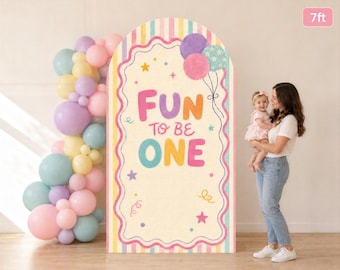 May include: A 7ft tall arched backdrop featuring a cream background with pastel vertical stripes, a scalloped pink border, and the text "FUN TO BE ONE" in colorful letters. It is decorated with illustrations of balloons and stars.