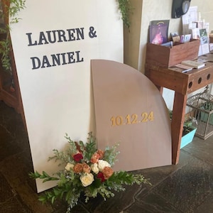 May include: A white and brown wedding sign with the text "Lauren & Daniel" and "10.12.24" in gold lettering. The sign is decorated with a bouquet of red, white, and orange roses.