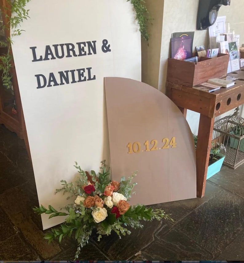May include: A white and brown wedding sign with the text "Lauren & Daniel" and "10.12.24" in gold lettering. The sign is decorated with a bouquet of red, white, and orange roses.
