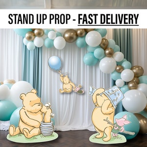 Puede incluir: Dos soportes de Winnie de Pooh de pie con un arco de globos en el fondo. Los soportes est&aacute;n hechos de cart&oacute;n y presentan a Winnie de Pooh en diferentes poses. El arco de globos est&aacute; hecho de globos blancos, dorados y azul claro.