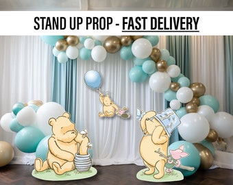 Op de afbeelding: Twee Winnie de Poeh stand-up rekwisieten met een ballonnenboog op de achtergrond. De rekwisieten zijn gemaakt van karton en tonen Winnie de Poeh in verschillende poses. De ballonnenboog is gemaakt van witte, gouden en lichtblauwe ballonnen.