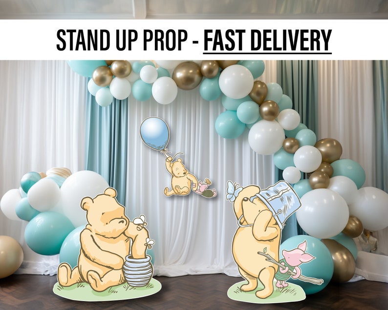 Puede incluir: Dos soportes de Winnie de Pooh de pie con un arco de globos en el fondo. Los soportes est&aacute;n hechos de cart&oacute;n y presentan a Winnie de Pooh en diferentes poses. El arco de globos est&aacute; hecho de globos blancos, dorados y azul claro.