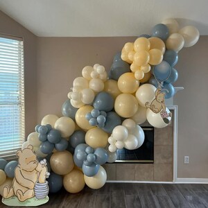 Puede incluir: Un arco de globos hecho de globos azules, blancos y beige. El arco est&aacute; decorado con personajes de Winnie the Pooh y Piglet. El arco est&aacute; instalado frente a una chimenea.