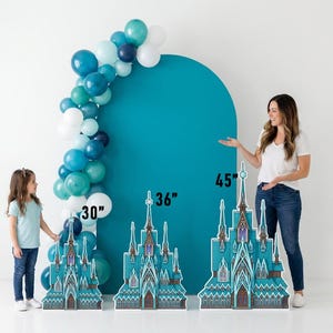 May include: A teal backdrop with a balloon arch and three castle cutouts. The castle cutouts are 30, 36, and 45 inches tall. The balloon arch features white, teal, and navy blue balloons. A woman and a child are standing near the cutouts.