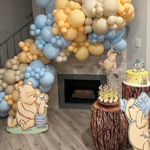 Puede incluir: Una fiesta de cumplea&ntilde;os con tem&aacute;tica de Winnie de Pooh con un arco de globos, un pastel y recortes de Winnie de Pooh. El pastel est&aacute; en un soporte de pastel de madera y los recortes est&aacute;n en el suelo.