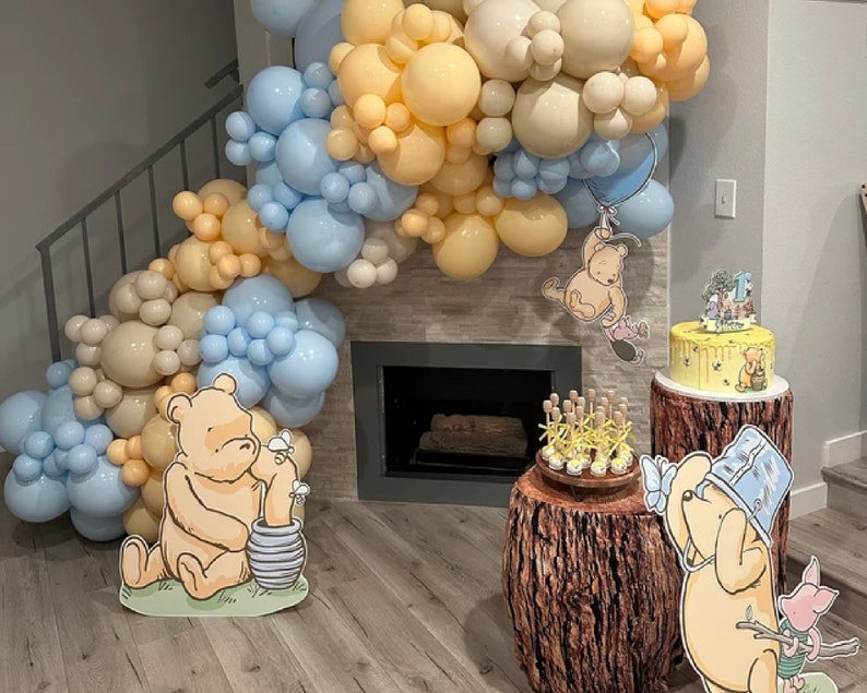 Puede incluir: Una fiesta de cumplea&ntilde;os con tem&aacute;tica de Winnie de Pooh con un arco de globos, un pastel y recortes de Winnie de Pooh. El pastel est&aacute; en un soporte de pastel de madera y los recortes est&aacute;n en el suelo.