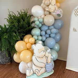 Puede incluir: Una decoraci&oacute;n de fiesta con tem&aacute;tica de Winnie the Pooh con un gran recorte de cart&oacute;n de Winnie the Pooh sosteniendo un tarro de miel. El fondo es un arco de globos con globos azules, verdes y beige.