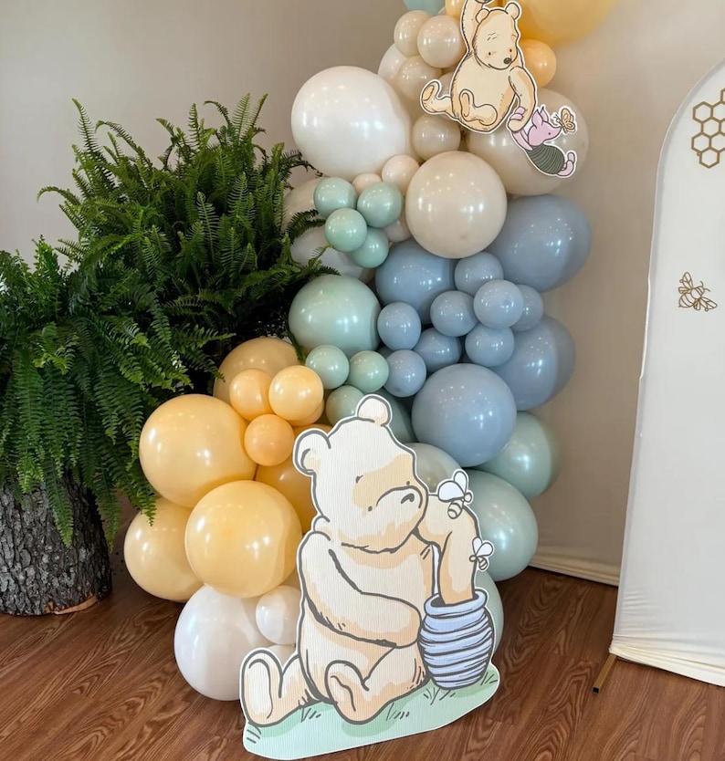 Puede incluir: Una decoraci&oacute;n de fiesta con tem&aacute;tica de Winnie the Pooh con un gran recorte de cart&oacute;n de Winnie the Pooh sosteniendo un tarro de miel. El fondo es un arco de globos con globos azules, verdes y beige.