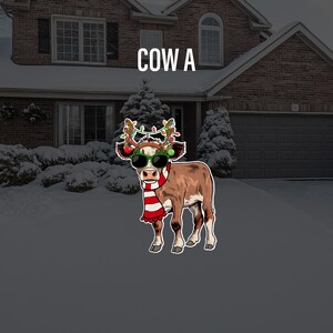 May include: A cartoon cow wearing sunglasses and a red and white scarf with Christmas lights on its antlers. The cow is standing in front of a snowy house with a garage door.