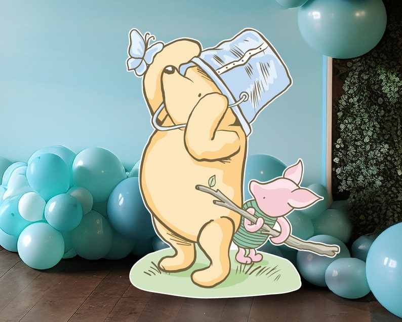 Puede incluir: Un recorte de Winnie de Pooh y Piglet con un cubo azul en la cabeza de Pooh. Pooh sostiene un palo y Piglet se agarra al palo. El recorte est&aacute; frente a una pared azul con una guirnalda de globos azules y blancos.