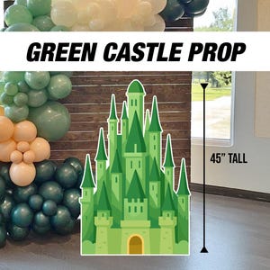 Może przedstawiać: Zielony rekwizyt zamku o wysokości 114 cm, z napisem "GREEN CASTLE PROP". Zamek to ilustracja z kreskówek z jasnozielonym korpusem i ciemniejszymi zielonymi akcentami. W tle łuk balonowy w odcieniach zieleni i brzoskwini.