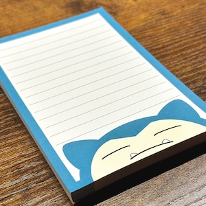 May include: A rectangular notepad with a blue border and horizontal lines for writing. The bottom section features a graphic of a sleeping blue and cream-colored creature with pointed ears and small teeth, positioned against a white background.