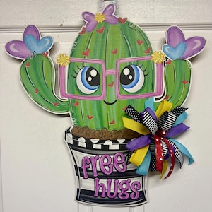 May include: A whimsical cactus-shaped wooden door hanger. The green cactus has a smiling face, pink glasses, and red heart accents. It is in a black and white striped pot with the words "free hugs". A colorful bow adds a decorative touch.