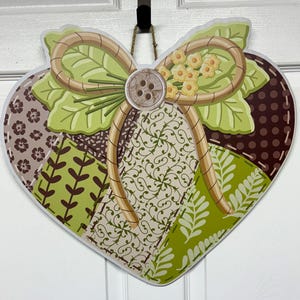 May include: A heart-shaped wall hanging featuring a patchwork design of green, brown, and cream fabric patterns. The center is decorated with a large tan ribbon bow, light green leaves, small yellow flowers, and a central brown four-hole button.