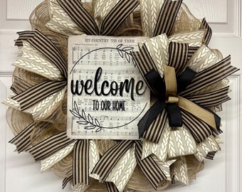 May include: A decorative wreath with a sign that reads "welcome to our home." The wreath features a mix of beige, brown, and black ribbons and mesh. The sign has musical notes and the words "My Country 'Tis of Thee."