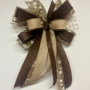 May include: A decorative bow featuring a mix of brown and tan ribbons. The bow has a variety of textures, including a patterned fabric with white cotton bolls, a textured solid brown ribbon, and a tan and white checkered ribbon. A rustic mesh adds a natural touch.