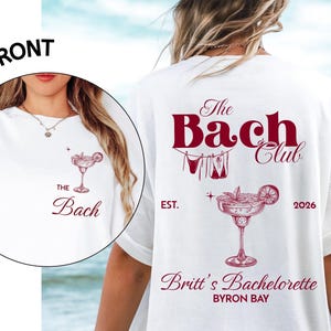May include: A white t-shirt featuring a maroon graphic design. The back displays the text The Bach Club, a margarita illustration, the text EST. 2026, Britt's Bachelorette, and BYRON BAY. The front pocket area shows a smaller margarita and The Bach.