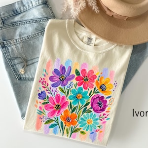 May include: An ivory t-shirt featuring a vibrant, multi-coloured floral graphic print. The design showcases various flowers in shades of purple, pink, orange, yellow, and teal, accented by painted brushstroke textures. The shirt has a crew neckline.