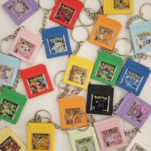 May include: A collection of colourful plastic keychains shaped like miniature Game Boy game cartridges, each featuring a unique Pokémon character label with the text Pokémon and Game Boy printed on the front of the rectangular casing.