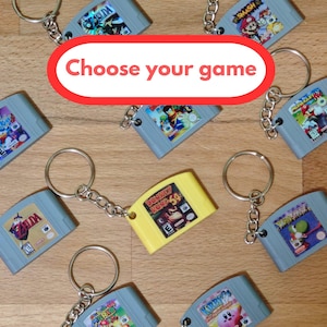 Puede incluir: Una colección de llaveros en miniatura con forma de cartuchos de videojuegos con títulos como Zelda, Mario Kart y Donkey Kong 64. Los llaveros son mayormente grises con un cartucho amarillo, unidos a una argolla de metal. El texto dice Choose your game.