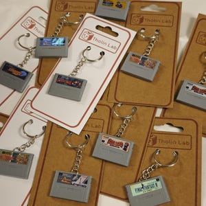 Peut inclure: Une collection de porte-clés miniatures en forme de cartouches de jeux vidéo gris, fixés à des anneaux et chaînes en métal. Chaque porte-clé est monté sur un support en carton rectangulaire avec le logo Tholin Lab et des titres de jeux.