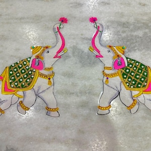 May include: Two decorative elephant figures with raised trunks holding pink flowers. The elephants are white with ornate green, gold, and pink detailing on their blankets and jewelry. The figures are likely for home decor.