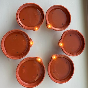 Puede incluir: Seis portavelas de té de color terracota con un borde decorativo y una pequeña luz redonda naranja en cada uno.