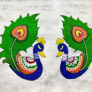 May include: Two colorful peacock designs. Each peacock has a blue body, yellow beak, and a green feather with an orange and yellow heart-shaped design. The background is a light gray marble.