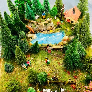 Può includere: Diorama in miniatura di una scena forestale con un fiume blu, alberi verdi e piccole figure di persone. Sono visibili anche una casa con il tetto rosso e una piccola mandria di mucche. La scena è ambientata su una base di legno.
