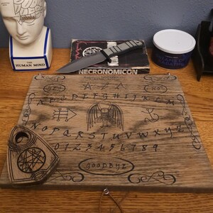 May include: A wooden Ouija board with a planchette, a skull, a book titled "Necronomicon", a knife, a ceramic head with a diagram of the human mind, and a pipe stand with two pipes.